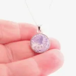 En hand håller i halsbandet Porslinssmycke - Lila Fasan, med ett silverhänge med en rund berlock som visar ett lila och vitt våginspirerat mönster.