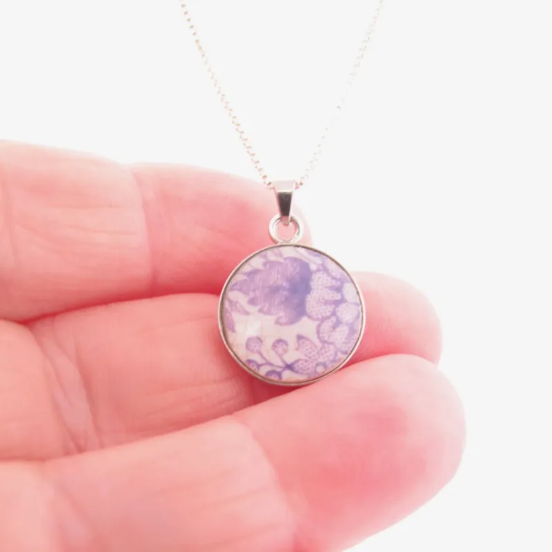 En hand håller i halsbandet Porslinssmycke - Lila Fasan, med ett silverhänge med en rund berlock som visar ett lila och vitt våginspirerat mönster.