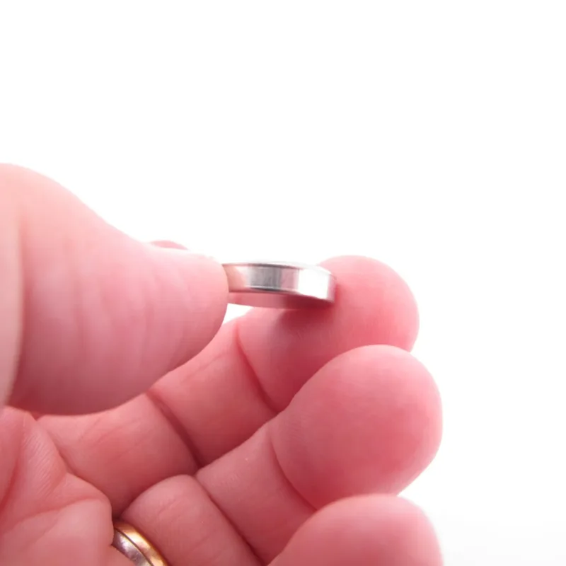 En hand håller Porslinsmycke - Lila Fasan, ett litet runt silverfärgat metallföremål, mellan tummen och pekfingret mot en vit bakgrund.