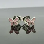 Ett par silverörhängen i form av fjärilar med vingar i rosa emalj, presenterade på en reflekterande yta.