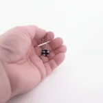 En hand håller i "Örhänge med porslin - Adam", ett enkelörhänge med en rund svartvit pärla med geometriskt mönster mot en vit bakgrund.