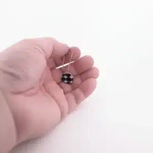 En hand håller i "Örhänge med porslin - Adam", ett enkelörhänge med en rund svartvit pärla med geometriskt mönster mot en vit bakgrund.