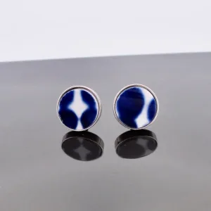 Ett par Örhänge med stift - Adam runda stiftörhängen med silverglänsande kant och blåvit abstrakt mitt, på en reflekterande grå yta.