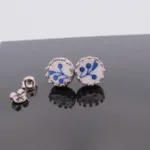 Örhängen med stift Krona - Dresden: Silverfärgade runda stiftörhängen med vit keramik i mitten och blått blommotiv. Det ena örhänget står upprätt, det andra ligger ner bredvid ett par örhängesbaksidor.