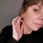 En person med ljus hy och ljusa ögon rör vid sitt Örhänge med stift - Dödskalle-örhänge, klädd i svart topp, bär en läppiercing i silver och en ring. Bakgrunden är enkel och neutral.