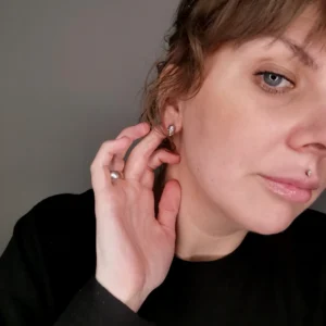 En person med ljus hy och ljusa ögon rör vid sitt Örhänge med stift - Dödskalle-örhänge, klädd i svart topp, bär en läppiercing i silver och en ring. Bakgrunden är enkel och neutral.