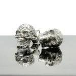 Två Örhänge med stift - Dödskalle, dödskalleformade örhängen i silver med intrikata detaljer, visas sida vid sida på en reflekterande yta mot en vit bakgrund.