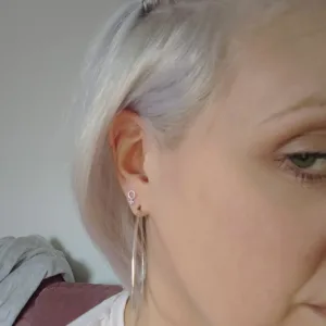 Närbild på en person med kort, ljusblont hår som bär Örhänge med stift - Kvinnosymbolen och en stor silverring. Endast en del av ansiktet är synligt, med en mjukt suddig bakgrund.