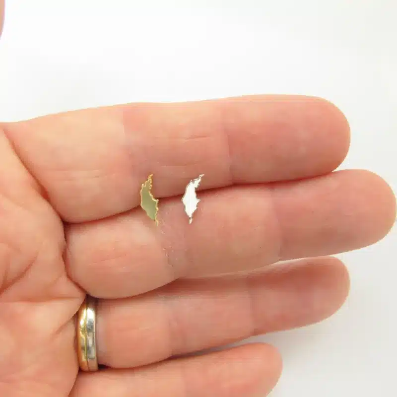 En hand med en guldring håller två små Örhängen med Stift - Gotland örhängen - ett guld och ett silver - på fingertopparna mot en vit bakgrund.