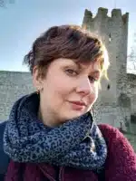 En kvinna med kort brunt hår och mönstrad halsduk, som bär Örhänge med Stift - Gotlands örhängen, ler lätt framför ett gammalt stentorn och en mur en solig dag.