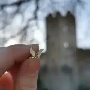 En hand håller upp Örhänge med Stift - Gotland, ett litet fladdermusformat örhänge i guld, framför en suddig bakgrund med ett stentorn och bladlösa trädgrenar.