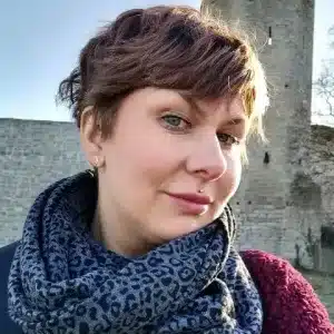En kvinna med kort brunt hår och mönstrad halsduk, som bär Örhänge med Stift - Gotlands örhängen, ler lätt framför ett gammalt stentorn och en mur en solig dag.