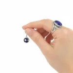 En hand med en stor blå stenring håller i "Örhänge Krona med porslin - Adam", ett litet runt örhänge med ett blåvitt stjärnmönster mot en vit bakgrund.