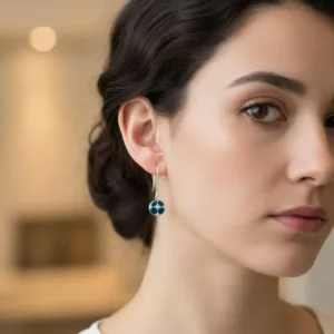 En kvinna med mörkt hår i en låg knut bär Örhänge med porslin - Adam, ett silverörhänge med ett blått cirkelformat hänge. Hon har ett lugnt ansiktsuttryck och bär en vit topp mot en mjukt suddig inomhusbakgrund.