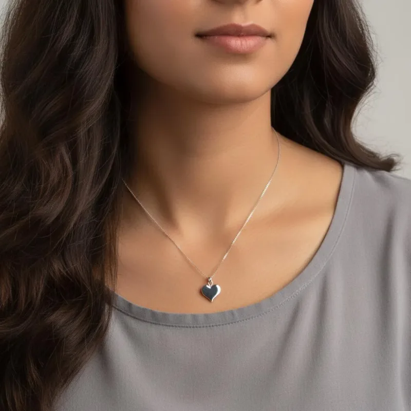 En kvinna med långt, vågigt brunt hår bär en ljusgrå topp och Handgjort Silverhjärta 27x15mm halsband. Bilden fångar hennes axlar upp, vilket framhäver silverhjärthänget och hennes subtila leende.