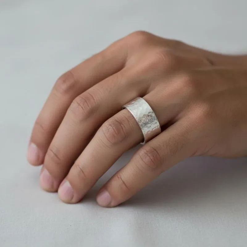 En hand med prydligt klippta naglar vilar på en ljus yta och visar upp Fingerring hamrad yta i äkta silver - en bred, hamrad silverring med en strukturerad, matt yta på långfingret.