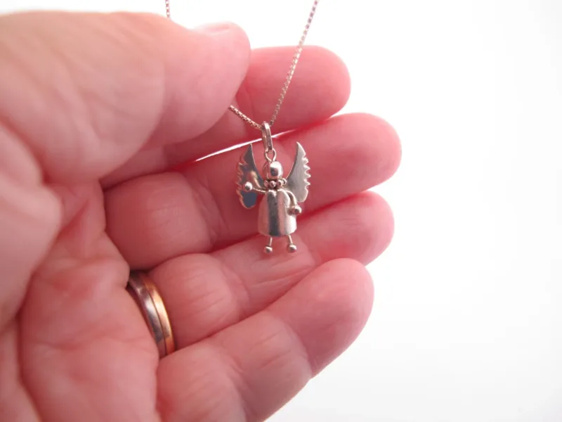 En hand håller i Handgjord Silverängel 26mm - en änglalik berlock i silver med vingar och runt huvud - på en kedja. Bakgrunden är vit och personen bär en guldring.