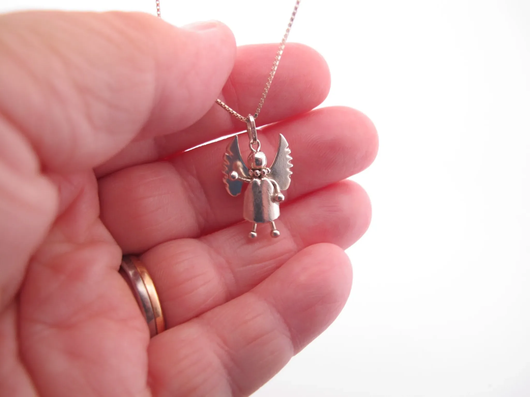 En hand håller i Handgjord Silverängel 26mm - en änglalik berlock i silver med vingar och runt huvud - på en kedja. Bakgrunden är vit och personen bär en guldring.