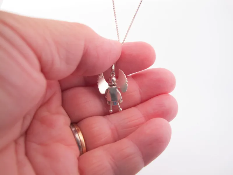 En hand håller i Handgjord Silverängel 26mm-halsbandet, ett änglahänge i silver med utsträckta vingar och enkel klädsel. Handen bär en vigselring i guld, infattad mot en vit bakgrund.