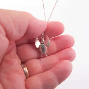 En hand håller i Handgjord Silverängel 26mm-halsbandet, ett änglahänge i silver med utsträckta vingar och enkel klädsel. Handen bär en vigselring i guld, infattad mot en vit bakgrund.