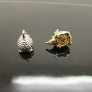 Två små, strukturerade metallberlocker - Silverberlock Igelkott 14 mm - formade som igelkottar visas på en reflekterande svart yta: en silverfärgad och upprätt, den andra guldfärgad och liggande på sidan med en synlig ihålig insida.
