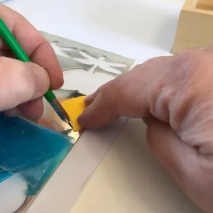 En närbild av händer som ritar en stencil på ett metallark för att skapa Silverplektrum Hajfena Handgjort Halsband, med hjälp av en grön penna och ett gult verktyg för att hålla stencilen på plats.
