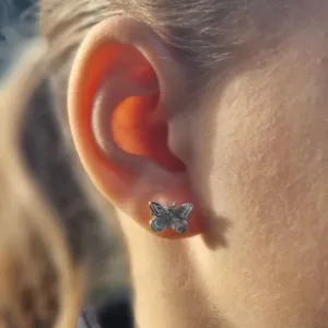 Närbild av en persons öra med Örhänge med stift - Fjäril, ett silverfärgat fjärilsformat stiftörhänge. Den mjukt suddiga bakgrunden framhäver örhängenas detaljerade design.