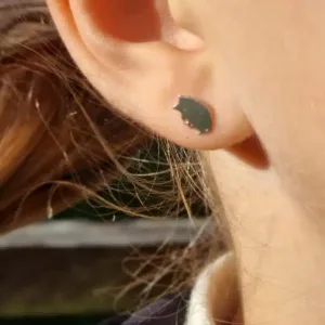 Närbild på en persons öra som bär "Örhänge med stift - Igelkott", ett litet runt stiftörhänge. Solljuset framhäver örat och det ljusbruna håret framträder mjukt i bakgrunden.