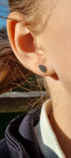 Närbild på en persons öra som bär "Örhänge med stift - Igelkott", ett litet runt stiftörhänge. Solljuset framhäver örat och det ljusbruna håret framträder mjukt i bakgrunden.