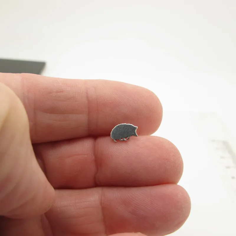 En person håller en Örhänge med stift - Igelkott, ett litet igelkottsformat örhänge i silver, på sina fingertoppar mot en vit bakgrund.
