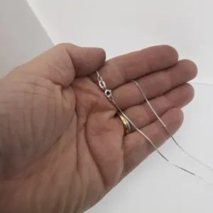 En hand med en guldring på ringfingret håller i Veneziakedja 0,8 mm Halskedja 42-45cm, en fin silverkedja med ett litet lås, mot en vit bakgrund.