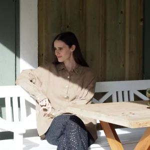 En kvinna med långt mörkt hår sitter på en vit bänk i solljus, klädd i GIA Linneskjorta en rak avslappnad modell i beige och svarta byxor med öljetter. Hon tittar åt sidan och vilar ena armen på ett träbord och den andra på bänken.