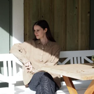 En kvinna med långt mörkt hår sitter på en vit bänk i solljus, klädd i GIA Linneskjorta en rak avslappnad modell i beige och svarta byxor med öljetter. Hon tittar åt sidan och vilar ena armen på ett träbord och den andra på bänken.