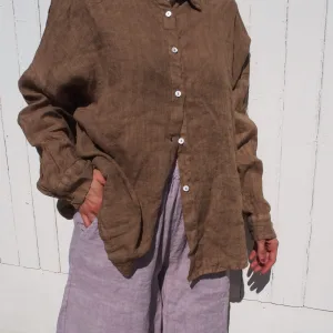En person står framför en vit trävägg, klädd i GIA Linneskjorta en rak avslappnad-modellen - en lös brun skjorta med knappar - och ljusgrå-lila shorts. En hand är i fickan, ansiktet är utanför bild.