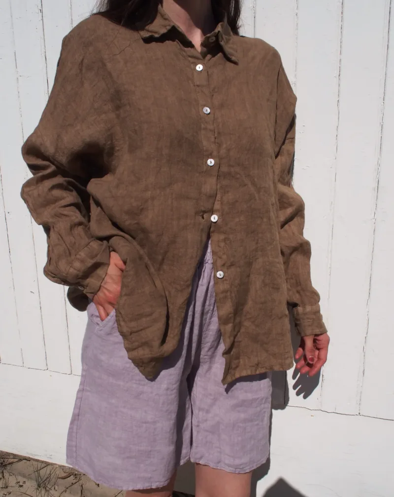 En person står framför en vit trävägg, klädd i GIA Linneskjorta en rak avslappnad-modellen - en lös brun skjorta med knappar - och ljusgrå-lila shorts. En hand är i fickan, ansiktet är utanför bild.