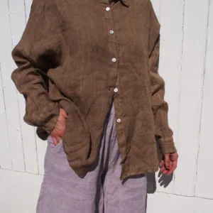En person står framför en vit trävägg, klädd i GIA Linneskjorta en rak avslappnad-modellen - en lös brun skjorta med knappar - och ljusgrå-lila shorts. En hand är i fickan, ansiktet är utanför bild.