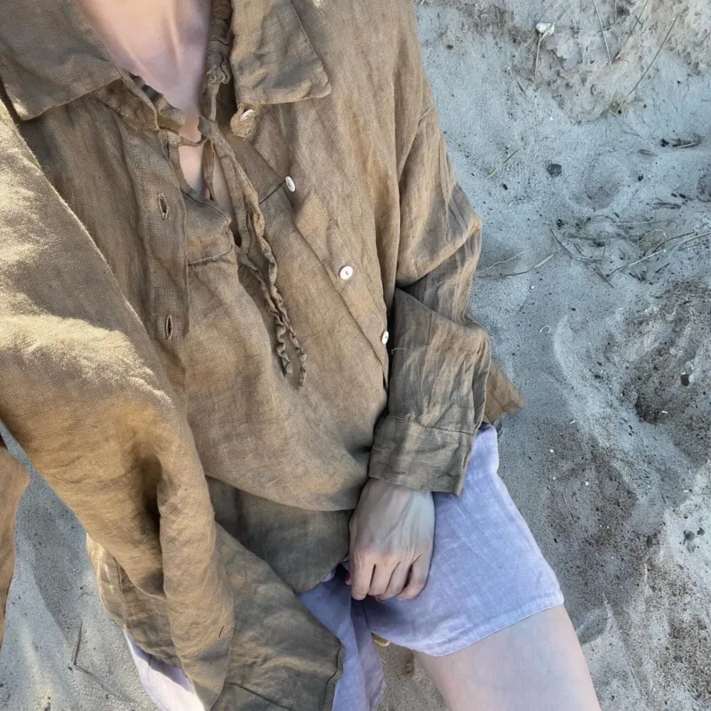 En person som bär GIA Linneskjorta, en brun långärmad skjorta med lös passform och ljusa shorts sitter på sandig mark i mjukt solljus. Huvudet är delvis utanför bild.