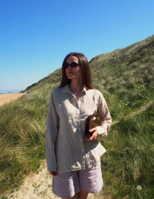 En kvinna i GIA Linneskjorta, en avslappnad beige linneskjorta, står på en solig gräsbevuxen sanddynsstig nära stranden med en bok under armen, blå himmel och gröna sanddyner syns bakom henne.