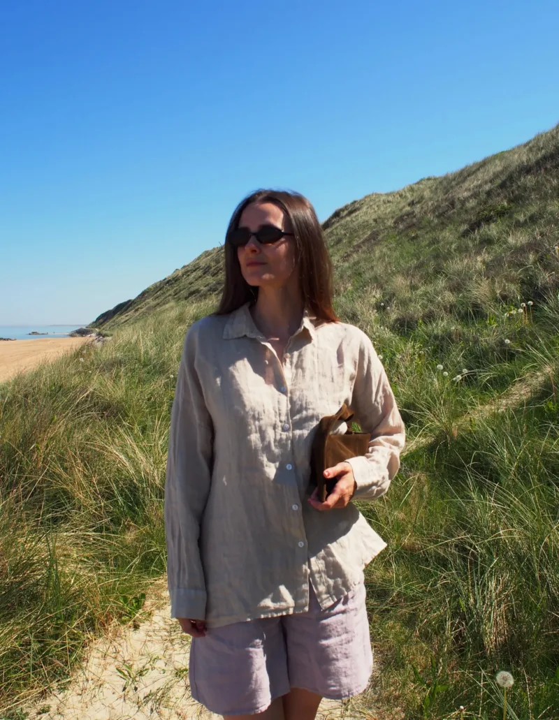 En kvinna i GIA Linneskjorta, en avslappnad beige linneskjorta, står på en solig gräsbevuxen sanddynsstig nära stranden med en bok under armen, blå himmel och gröna sanddyner syns bakom henne.