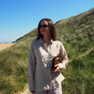 En kvinna i GIA Linneskjorta, en avslappnad beige linneskjorta, står på en solig gräsbevuxen sanddynsstig nära stranden med en bok under armen, blå himmel och gröna sanddyner syns bakom henne.