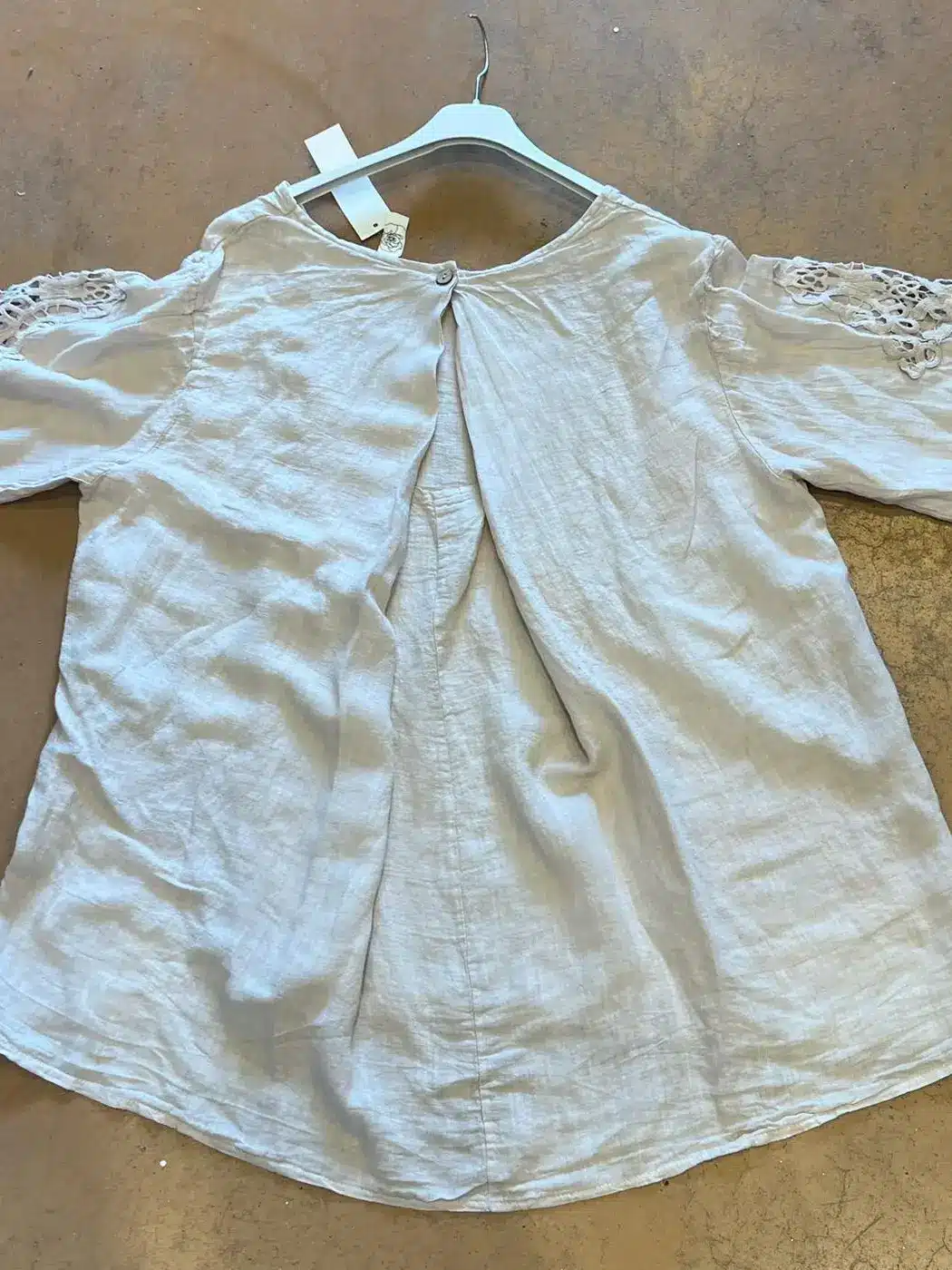 Light, loose linen blouse on a hanger with lace cutouts along the sleeves and a center back pleat with a small button at the neckline.