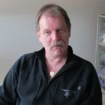 En medelålders man med ljust hår och mustasch sitter inomhus, klädd i en svart jacka med dragkedja och broderier och ett hjärtformat halsband med hänge, och tittar rakt in i kameran med ett neutralt ansiktsuttryck.