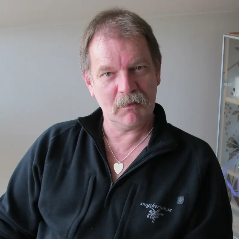 En medelålders man med ljust hår och mustasch sitter inomhus, klädd i en svart jacka med dragkedja och broderier och ett hjärtformat halsband med hänge, och tittar rakt in i kameran med ett neutralt ansiktsuttryck.