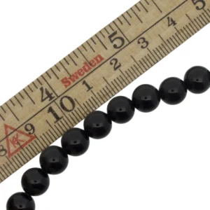 Halsbandet Svart rund onyx eller agat 4-10 mm är placerat bredvid en trälinjal med centimeter- och millimetermärken, vilket indikerar att varje pärla mäter cirka 1 centimeter i diameter.