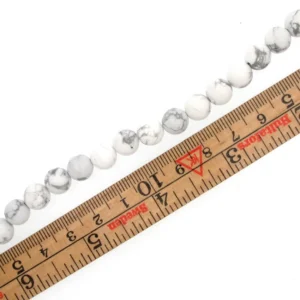 Howlit 8mm runda ädelstenspärlor runda och vita med grå marmorering, visas ovanför en trälinjal som visar centimeter och millimeter. Varje pärla mäter ca 8 mm i diameter.