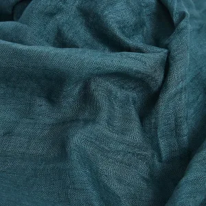 Närbild av texturerat tealfärgat tyg med synlig väv och naturliga veck som skapar mjuka skuggor och ljuspunkter.
