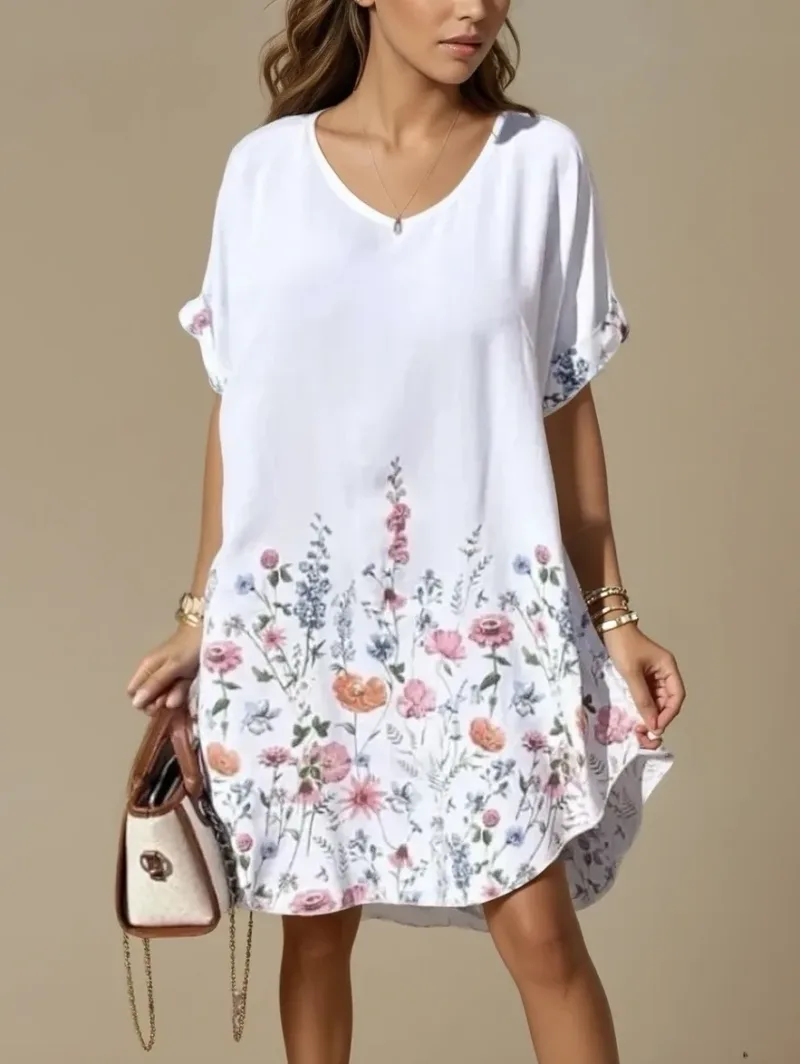 En kvinna bär Midsommartunikan/klänningen - en vit, kortärmad klänning med löst sittande passform och rosa, orange och blått blommönster nedtill - tillsammans med en brun handväska, armband och ett halsband.