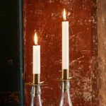 Två tända vita ljus i olika höjd i Ljushållare för Flaska i mässing ger ett varmt sken mot en strukturerad, rödbrun vägg.