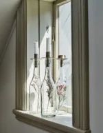 Två höga vita ljus sitter i Ljushållare för Flaska ovanpå glasflaskor med torkade blommor, placerade på en fönsterbräda där solljuset flödar in.