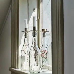 Två höga vita ljus sitter i Ljushållare för Flaska ovanpå glasflaskor med torkade blommor, placerade på en fönsterbräda där solljuset flödar in.